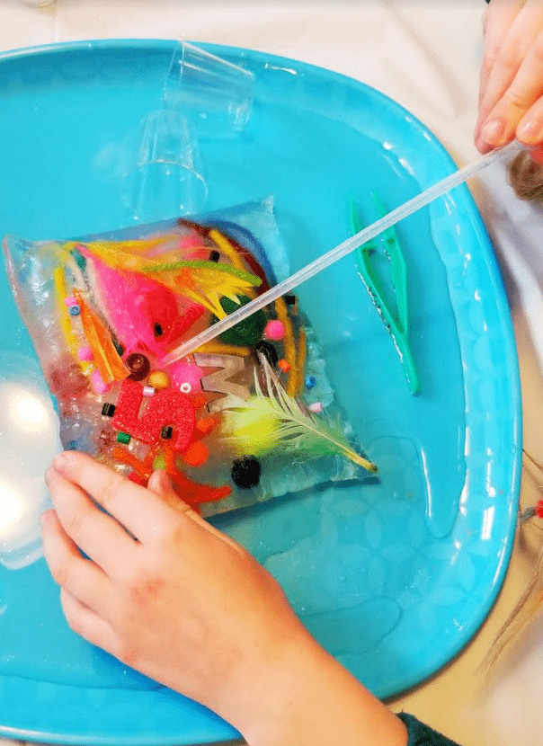frozen science experiment shows a child dropping water on an ice brick filled with craft supplies.