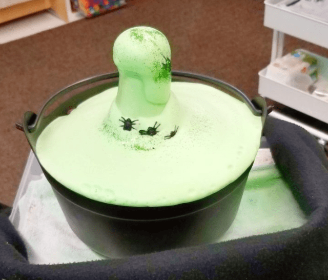 science experiment for kids shows a bowl like a witches cauldron with green foam coming out of it and plastic spiders.