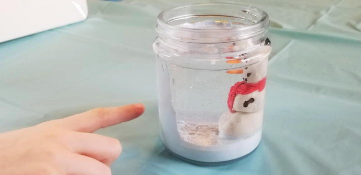 A child is pointing at their jar that holds their clay snowman filled with oil and paint falling like snow for this stem activity for kids