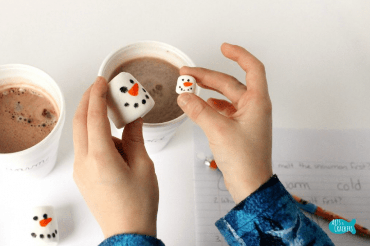 Christmas STEM activities shows a child holding a large and small marshmallow that looks like a snowman over hot chocolate.