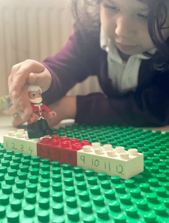 Math activity shows a child holding a Santa lego figure moving him over lego pieces with numbers.