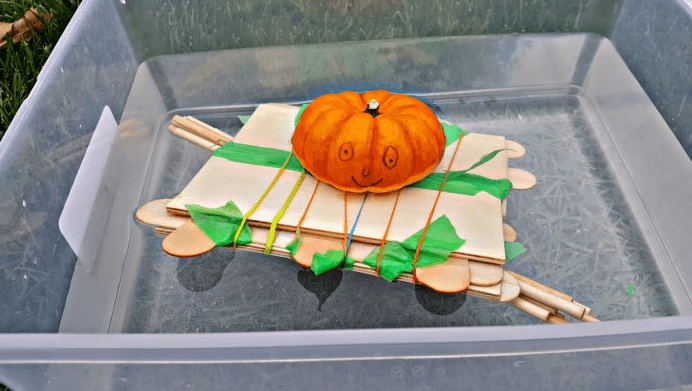 STEM challenge shows a mini pumpkin on a boat made from sticks, boards, elastics and tape.
