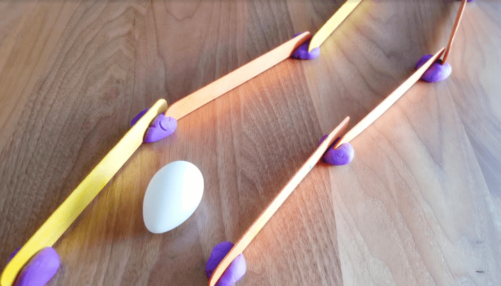 stem activity for kids shows popsicle sticks on a table attached with play dough and an egg in the path.