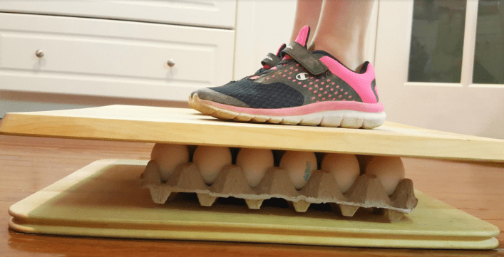egg experiments for kids shows a child standing on a board on top of a carton of eggs.