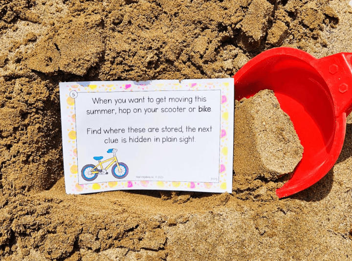 outdoor scavenger hunt shows a printed clue in the sand with a red shovel beside it.