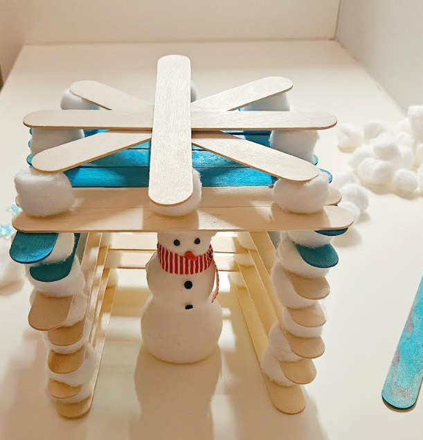 winter activity for kids shows a snowman toy inside of a home made from popsicle sticks and cotton balls,.